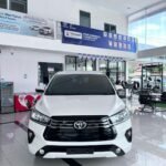 Front view of a white Toyota SUV on a showroom floor, bold black grille and emblem visible, with banners and glass doors in the background.