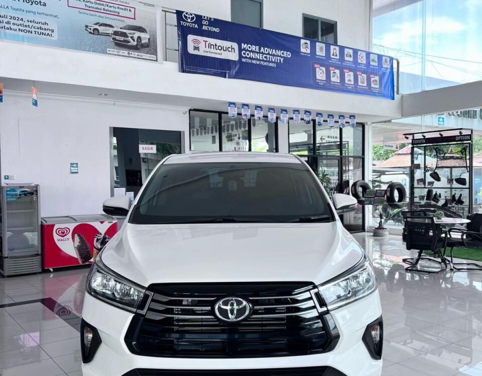 Front view of a white Toyota SUV on a showroom floor, bold black grille and emblem visible, with banners and glass doors in the background.