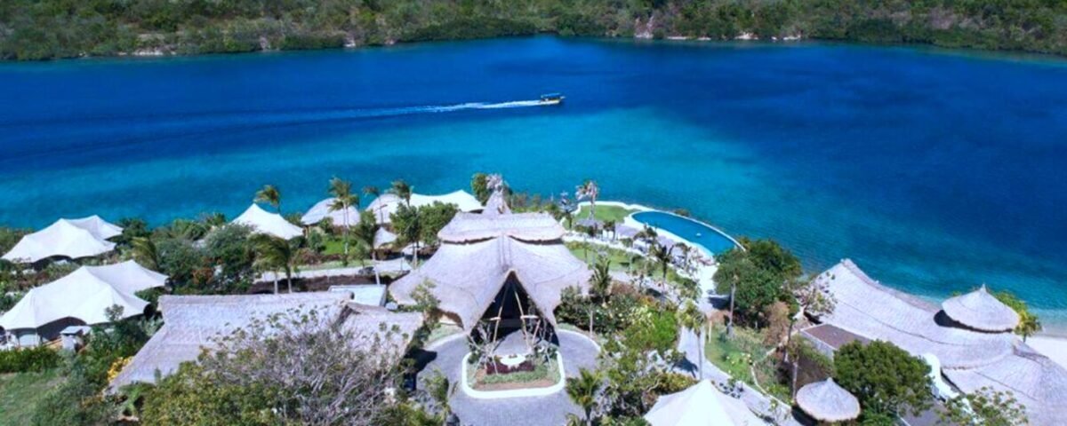 A tropical resort with thatched-roof villas around a blue lagoon and a boat cruising the water.
