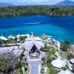 A tropical resort with thatched-roof villas around a blue lagoon and a boat cruising the water.