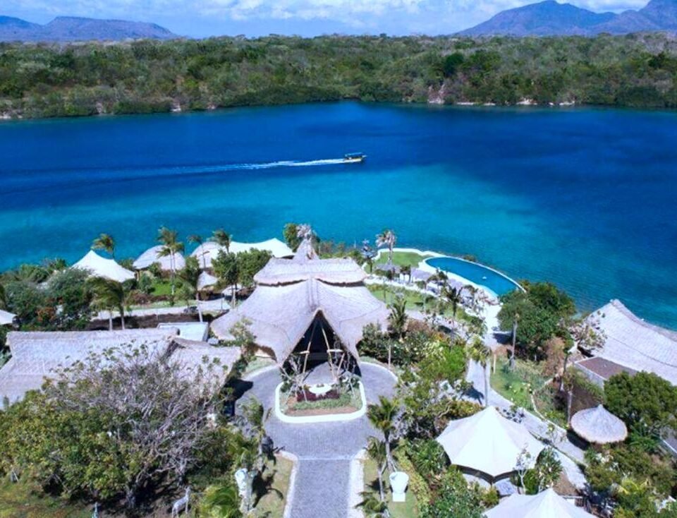 A tropical resort with thatched-roof villas around a blue lagoon and a boat cruising the water.