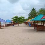 Row of colorful beach pavilions with benches along a sandy shore, palm trees in the background under a cloudy sky.