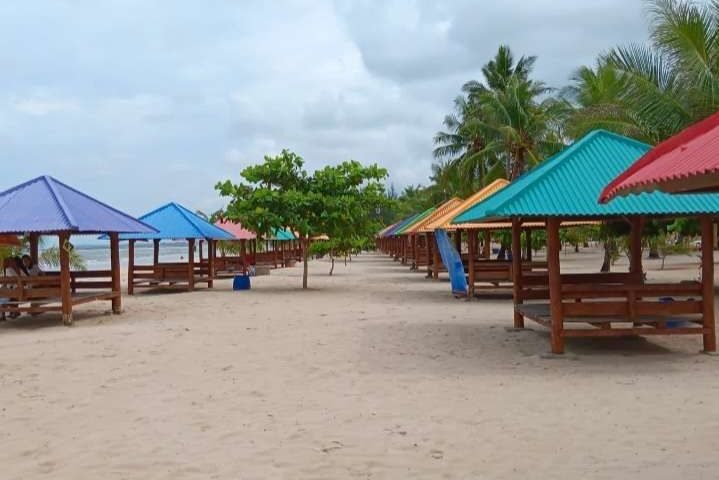Row of colorful beach pavilions with benches along a sandy shore, palm trees in the background under a cloudy sky.