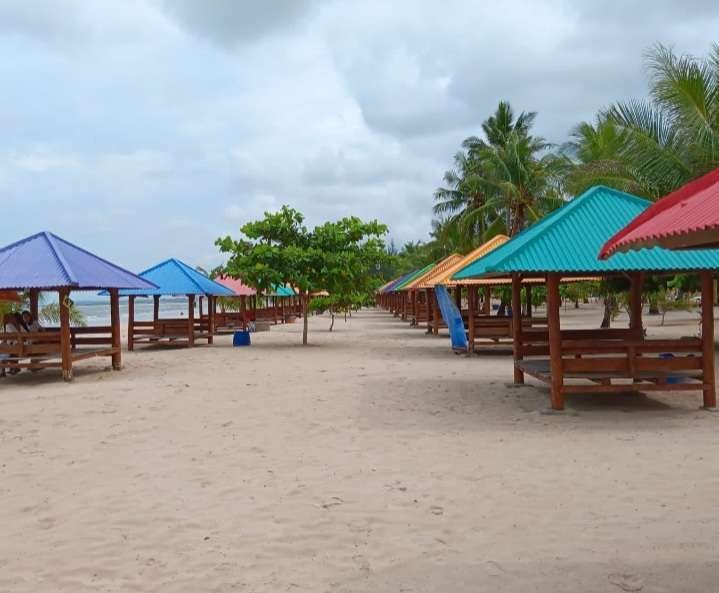 Row of colorful beach pavilions with benches along a sandy shore, palm trees in the background under a cloudy sky.