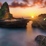 Sunset over calm sea with rocky formations; a person stands on a rock near a small tent on the right ground edge.