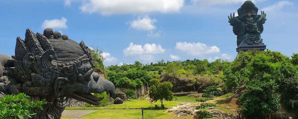 Giant dark stone dragon head sculpture in a sunny park, with a towering multi-armed statue on a hill in the background.
