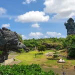 Giant dark stone dragon head sculpture in a sunny park, with a towering multi-armed statue on a hill in the background.
