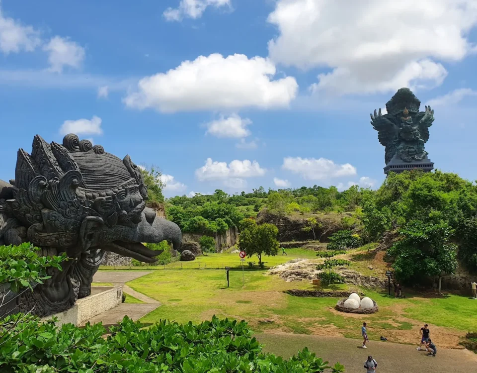 Giant dark stone dragon head sculpture in a sunny park, with a towering multi-armed statue on a hill in the background.