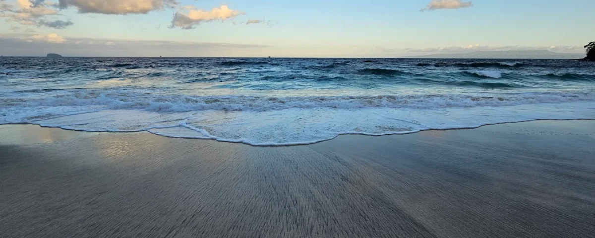 Blue ocean with small waves reaching a smooth sandy shore under a clear sky with a few clouds. (beach scene)