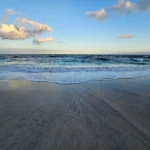 Blue ocean with small waves reaching a smooth sandy shore under a clear sky with a few clouds. (beach scene)