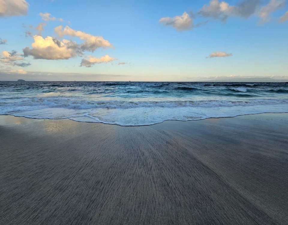 Blue ocean with small waves reaching a smooth sandy shore under a clear sky with a few clouds. (beach scene)