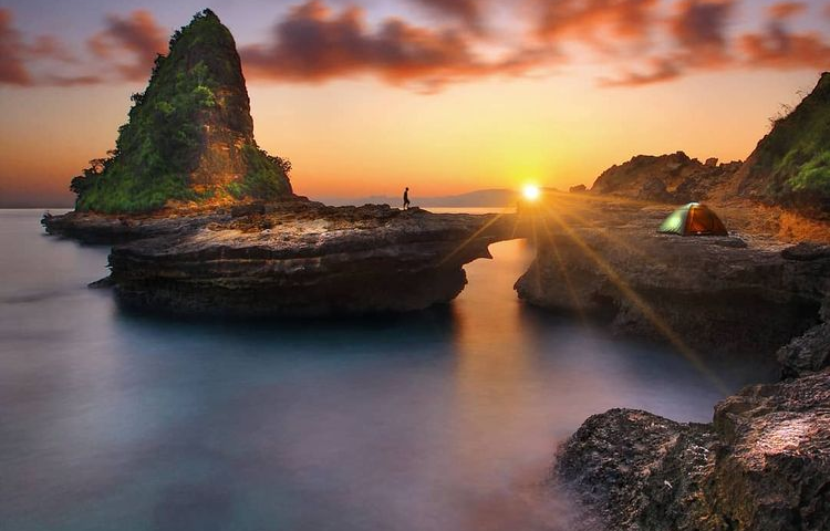 Sunset over calm sea with rocky formations; a person stands on a rock near a small tent on the right ground edge.