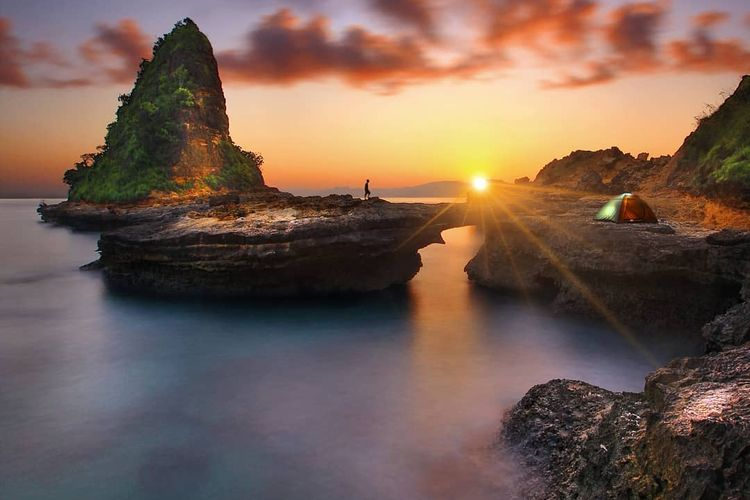 Sunset over calm sea with rocky formations; a person stands on a rock near a small tent on the right ground edge.
