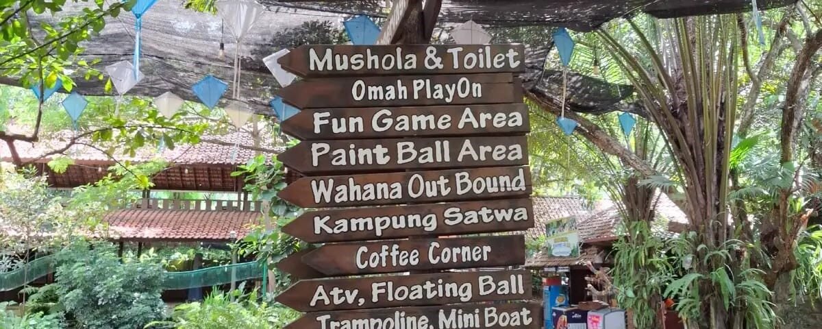 Wooden signpost in a lush outdoor recreation area listing amenities like Mushola & Toilet, Paint Ball, Fun Game Area, and Coffee Corner.