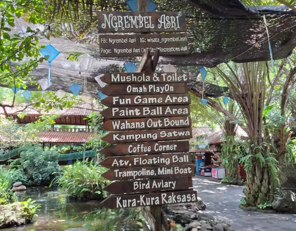 Wooden signpost in a lush outdoor recreation area listing amenities like Mushola & Toilet, Paint Ball, Fun Game Area, and Coffee Corner.