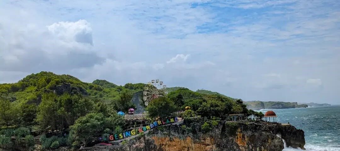 Rocky coastal outcrop with turquoise waves crashing below; greenery on top and a colorful sign along the edge.