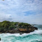Rocky coastal outcrop with turquoise waves crashing below; greenery on top and a colorful sign along the edge.