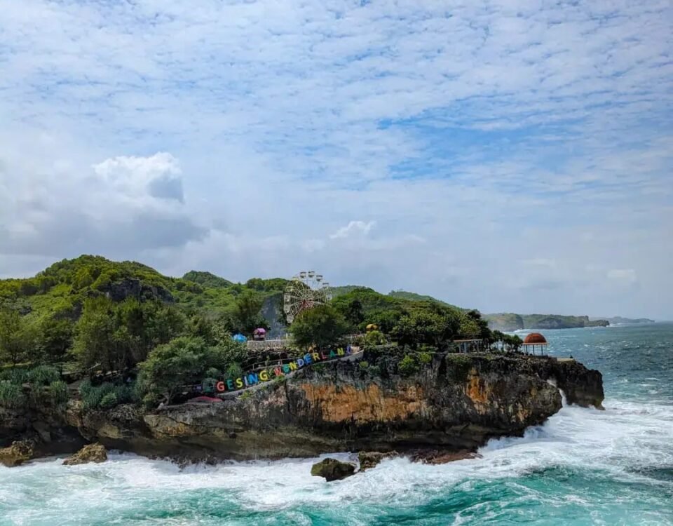 Rocky coastal outcrop with turquoise waves crashing below; greenery on top and a colorful sign along the edge.