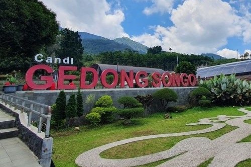 Large red sign reading 'GEDONG SONGO' on a landscaped hillside with stairs and distant mountains under a blue, partly cloudy sky.