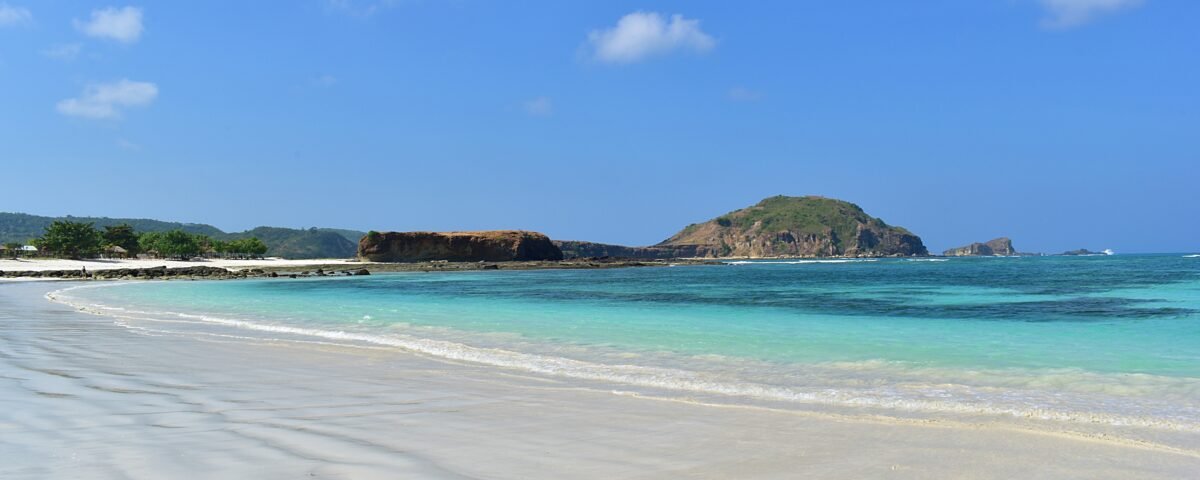 Tropical beach with white sand, turquoise water, and a rocky island on the horizon under a blue sky.