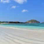Tropical beach with white sand, turquoise water, and a rocky island on the horizon under a blue sky.