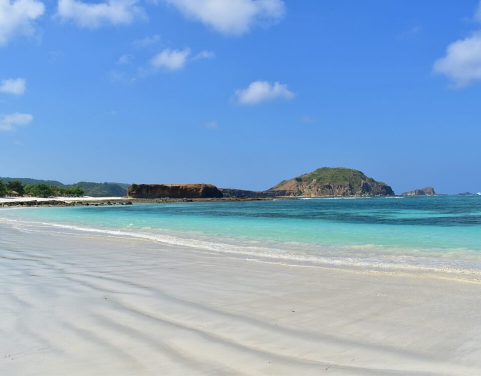 Tropical beach with white sand, turquoise water, and a rocky island on the horizon under a blue sky.