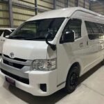 White Toyota van in a showroom, front three-quarter view, clean and polished interior lighting reflecting off the body