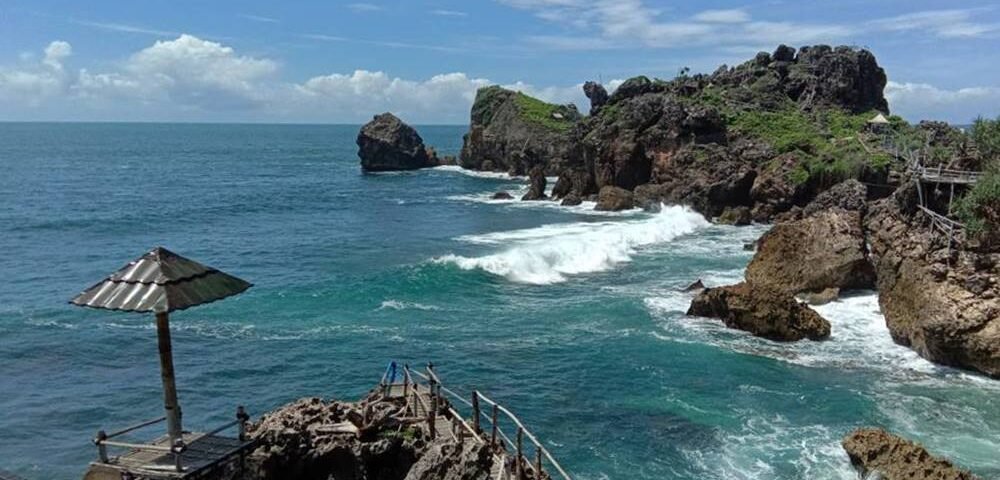 Rocky coastline with turquoise waves, a wooden viewing platform, and a thatched-roof shelter on the left.