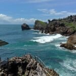Rocky coastline with turquoise waves, a wooden viewing platform, and a thatched-roof shelter on the left.