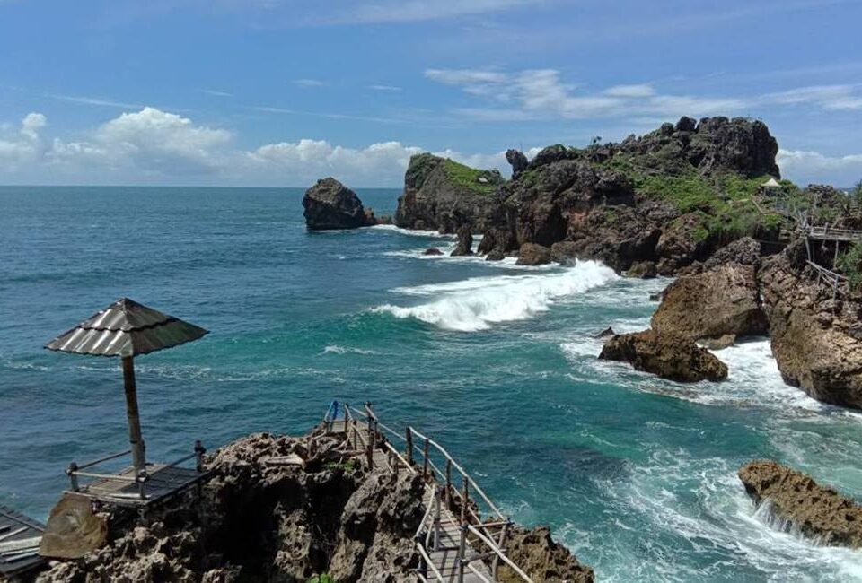 Rocky coastline with turquoise waves, a wooden viewing platform, and a thatched-roof shelter on the left.
