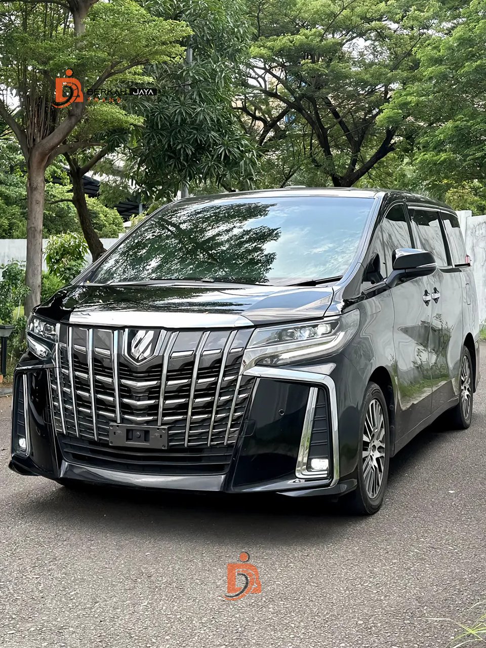 Front three-quarter view of a dark gray luxury minivan with a bold chrome grille, parked on a tree-lined street.