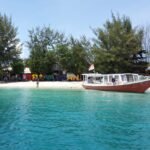 Turquoise water near a white-sand beach with a red and white tour boat moored at the shore, and colorful letters spelling a sign behind the trees.