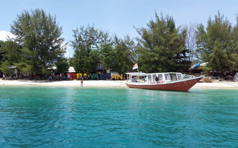 Turquoise water near a white-sand beach with a red and white tour boat moored at the shore, and colorful letters spelling a sign behind the trees.