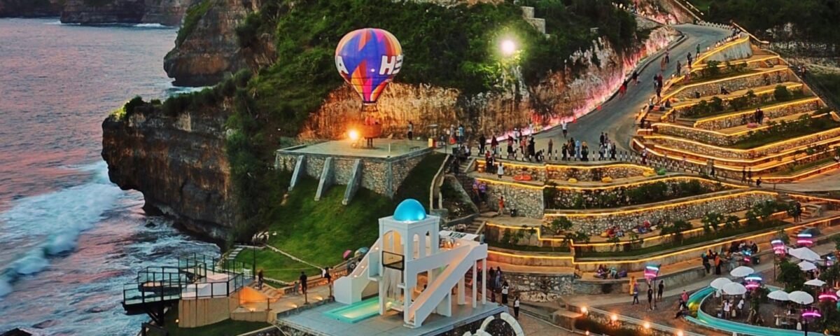 Sunset over a cliffside coastal park with illuminated terraced walkways, a colorful hot air balloon near a launch platform, and visitors strolling the paths along the cliff edge by the sea.