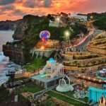 Sunset over a cliffside coastal park with illuminated terraced walkways, a colorful hot air balloon near a launch platform, and visitors strolling the paths along the cliff edge by the sea.