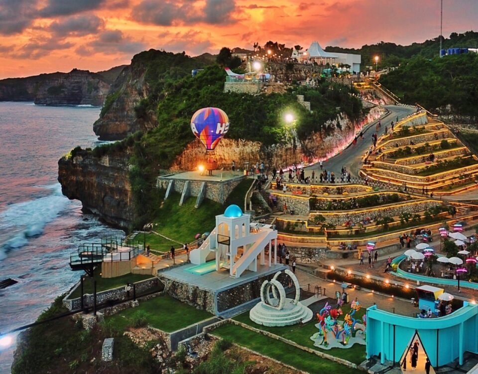Sunset over a cliffside coastal park with illuminated terraced walkways, a colorful hot air balloon near a launch platform, and visitors strolling the paths along the cliff edge by the sea.