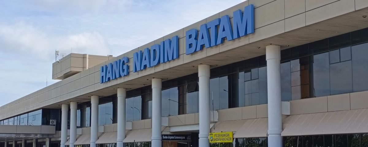 Exterior view of Hang Nadim Batam airport terminal with a large blue sign, cars parked in front, and orange traffic cones in the foreground.