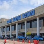 Exterior view of Hang Nadim Batam airport terminal with a large blue sign, cars parked in front, and orange traffic cones in the foreground.