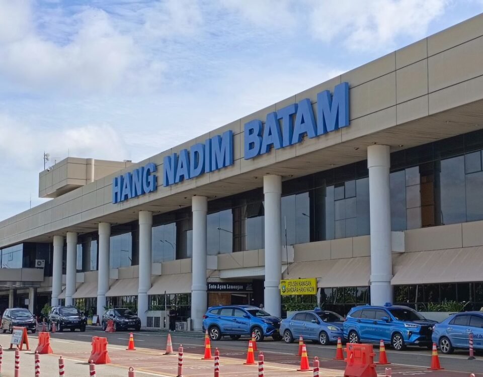 Exterior view of Hang Nadim Batam airport terminal with a large blue sign, cars parked in front, and orange traffic cones in the foreground.