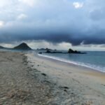 Desolate beach with wet sand and calm water under a looming dark storm cloud over distant rocky headlands