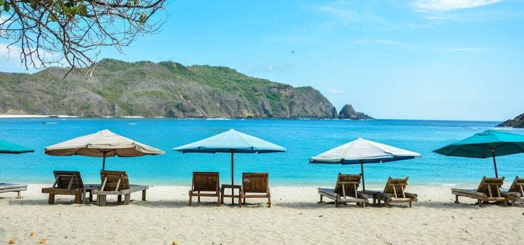 Sunlit beach with wooden lounge chairs under blue and turquoise umbrellas, calm sea and distant green hills.