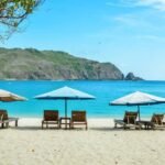 Sunlit beach with wooden lounge chairs under blue and turquoise umbrellas, calm sea and distant green hills.