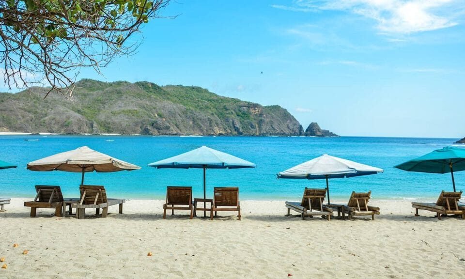 Sunlit beach with wooden lounge chairs under blue and turquoise umbrellas, calm sea and distant green hills.
