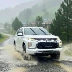White SUV splashing through a shallow puddle on a gravel rural road with green hills and trees in the misty background.