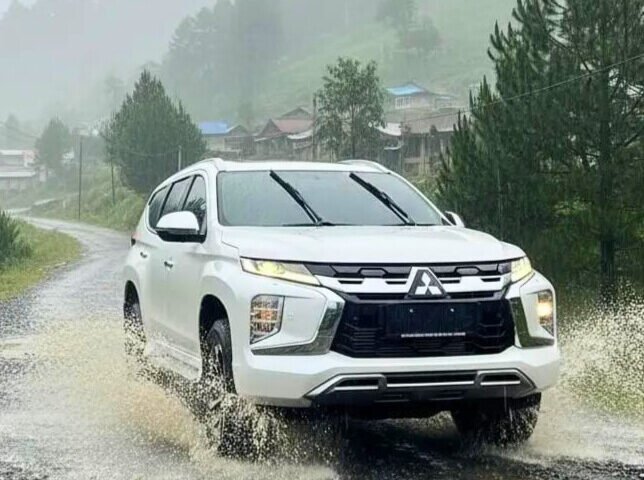 White SUV splashing through a shallow puddle on a gravel rural road with green hills and trees in the misty background.