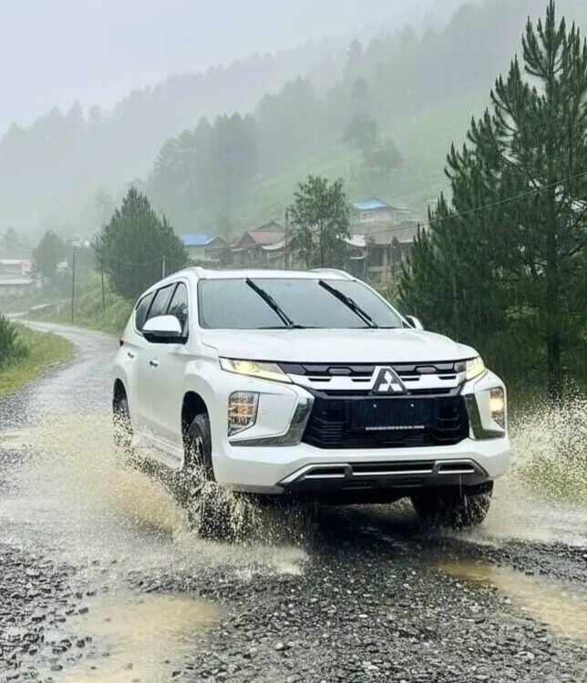 White SUV splashing through a shallow puddle on a gravel rural road with green hills and trees in the misty background.