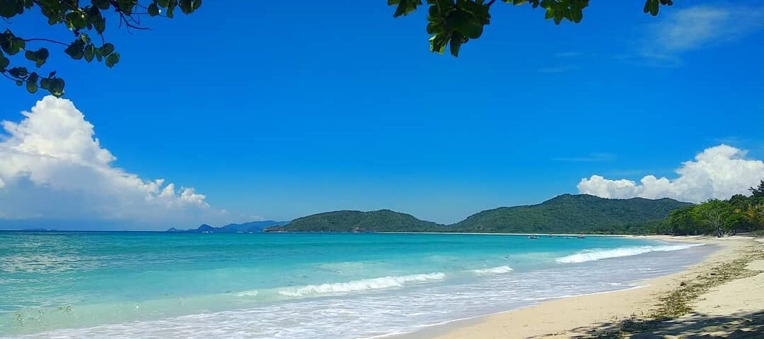 Sunny tropical beach with white sand, turquoise water, and distant green hills framed by leafy branches overhead.