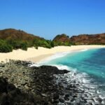 Tropical beach with white sand, turquoise water, and rocky foreground backed by green shrubs and hills under a clear blue sky.