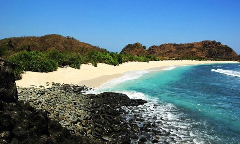 Tropical beach with white sand, turquoise water, and rocky foreground backed by green shrubs and hills under a clear blue sky.