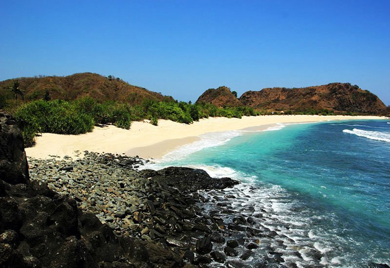 Tropical beach with white sand, turquoise water, and rocky foreground backed by green shrubs and hills under a clear blue sky.